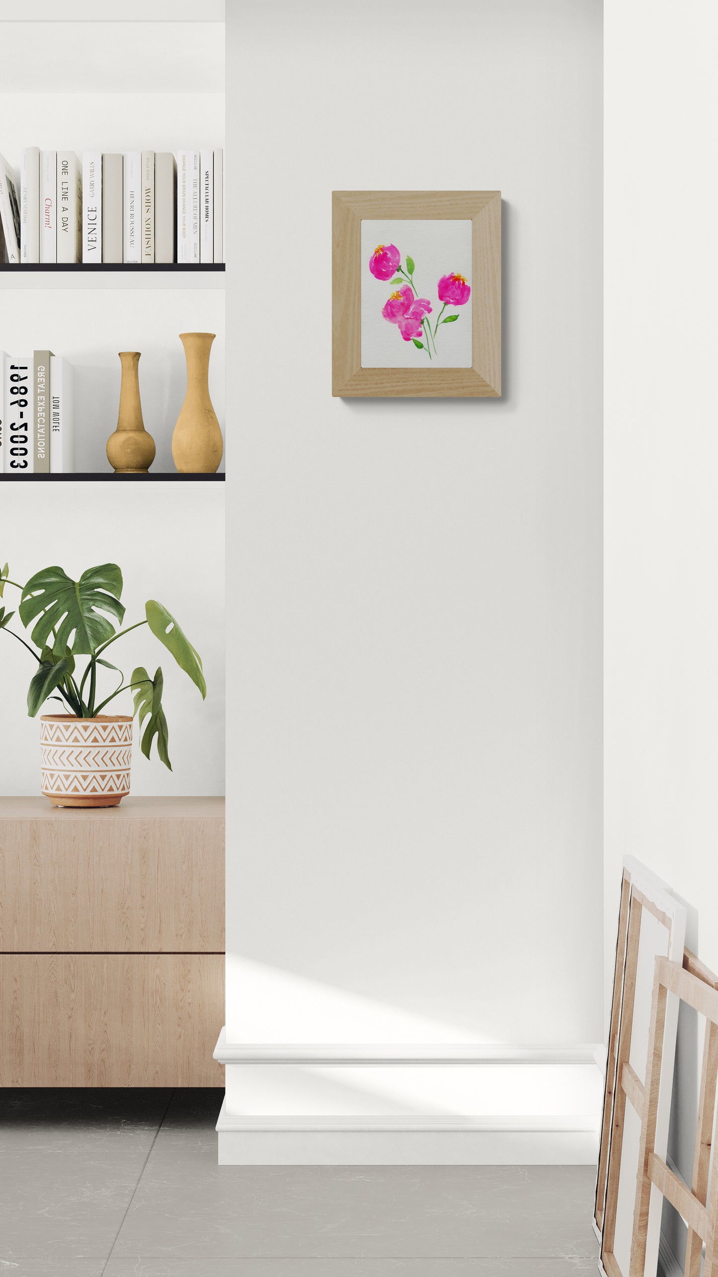 Framed artwork of pink flowers on a white wall above a wooden cabinet with a plant.