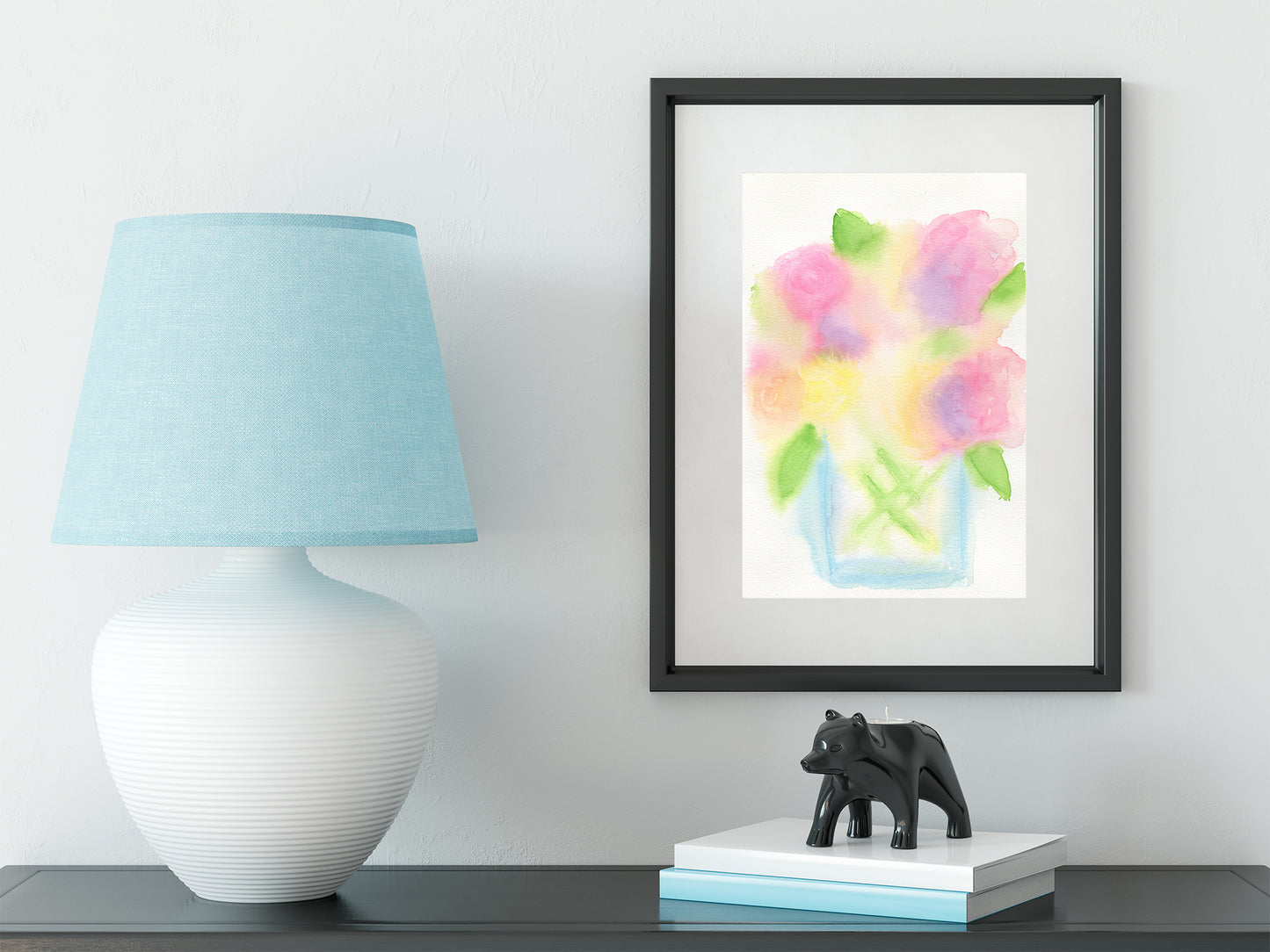 Framed watercolor painting of flowers on a wall above a table with a lamp and decorative items.