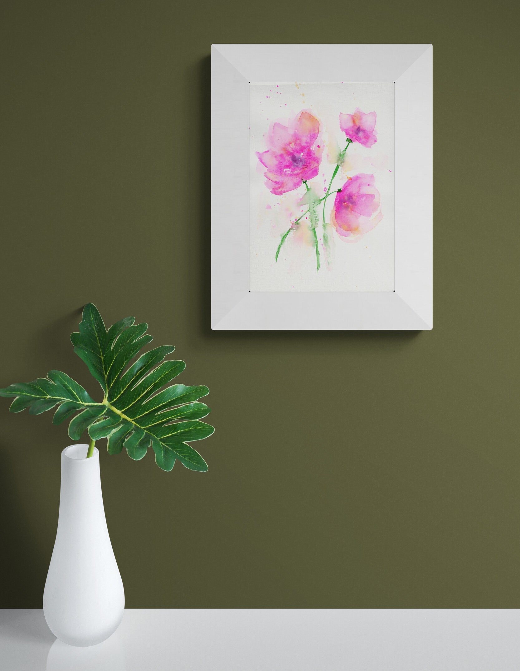 Framed artwork of pink flowers on a green wall with a white vase and leaf in the foreground.