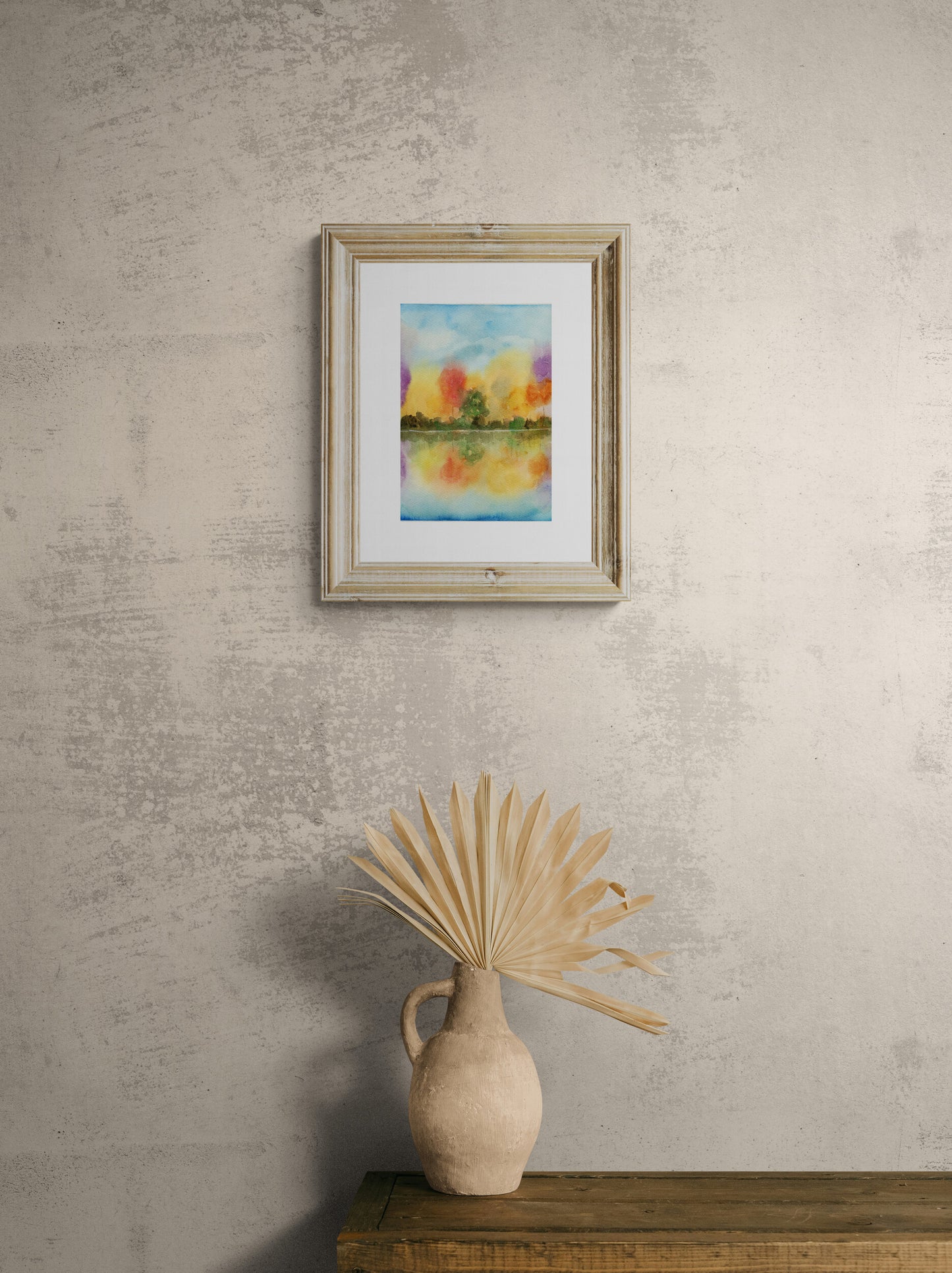 Framed artwork on a textured wall with a vase and dried plant on a wooden surface below.