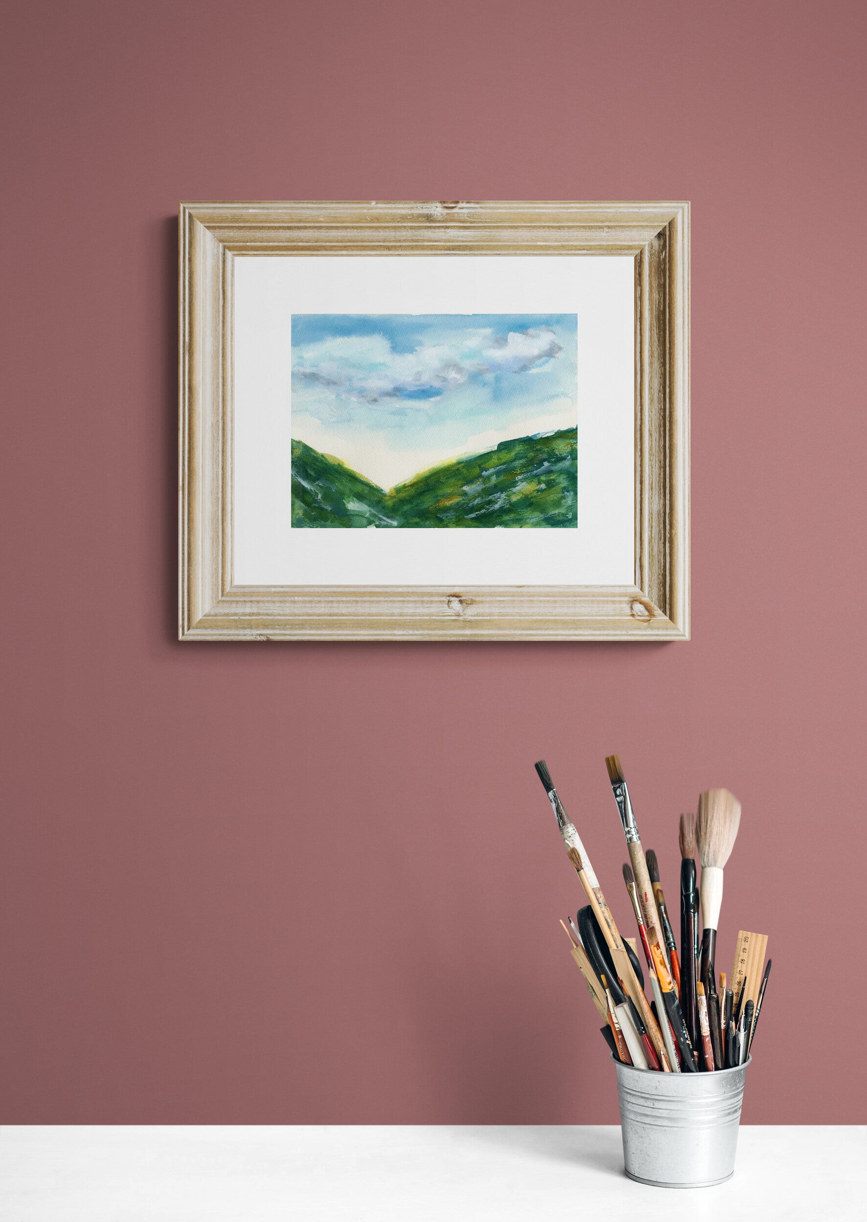 Framed painting of a landscape on a pink wall with paintbrushes in a container below.