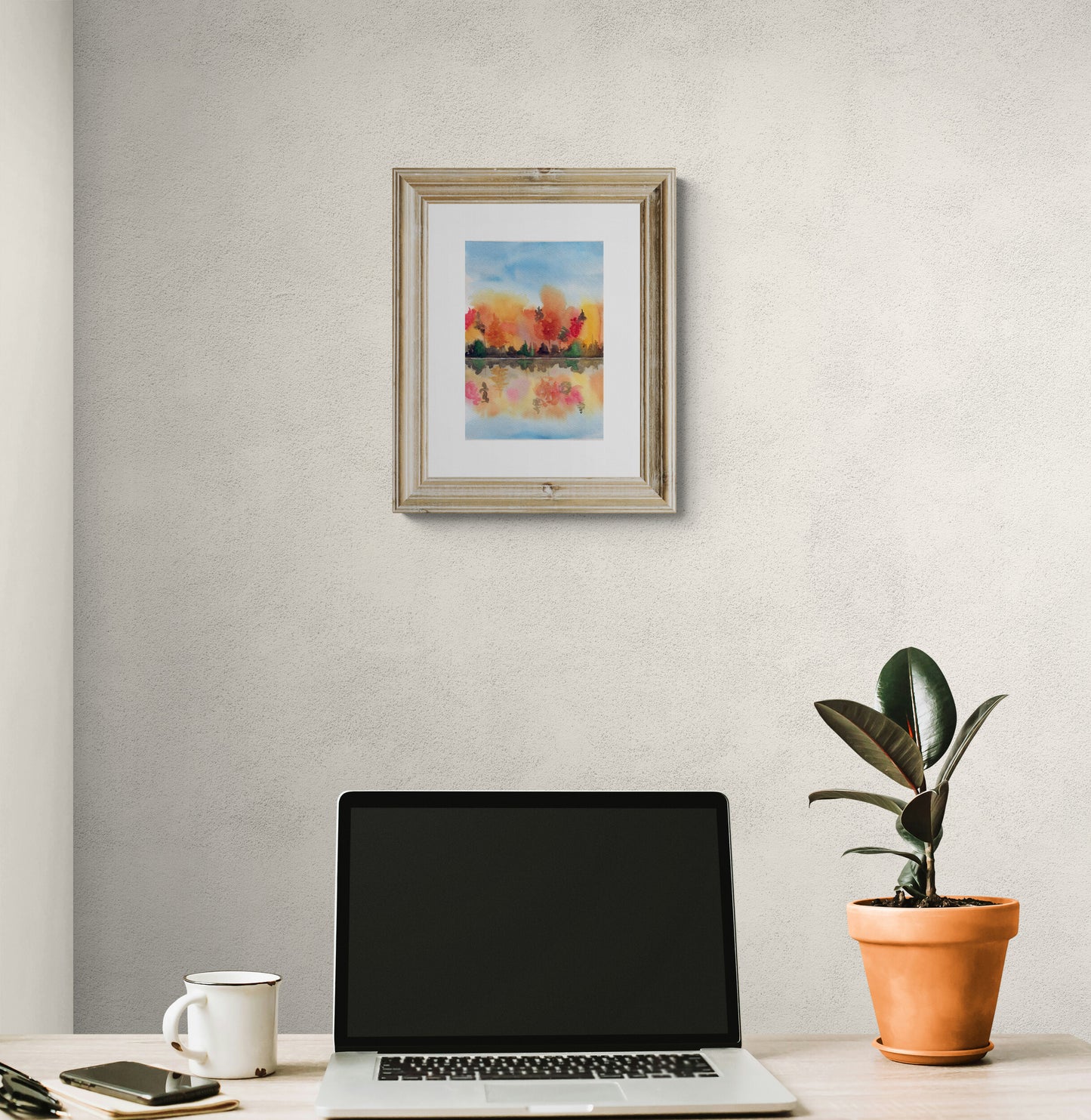 Framed artwork above a laptop on a desk with a plant and mug.
