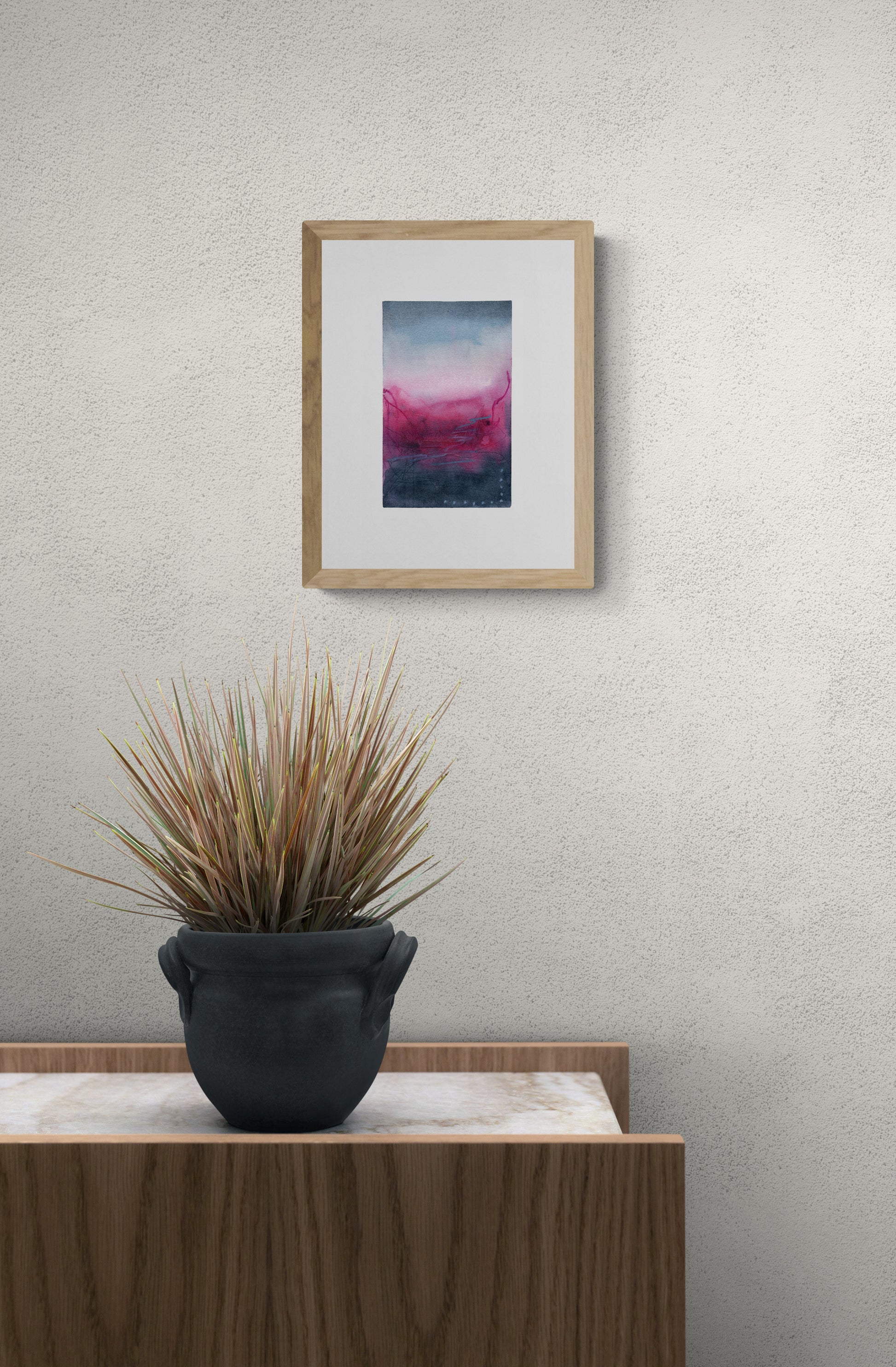 Framed abstract art on a wall above a potted plant on a wooden surface.