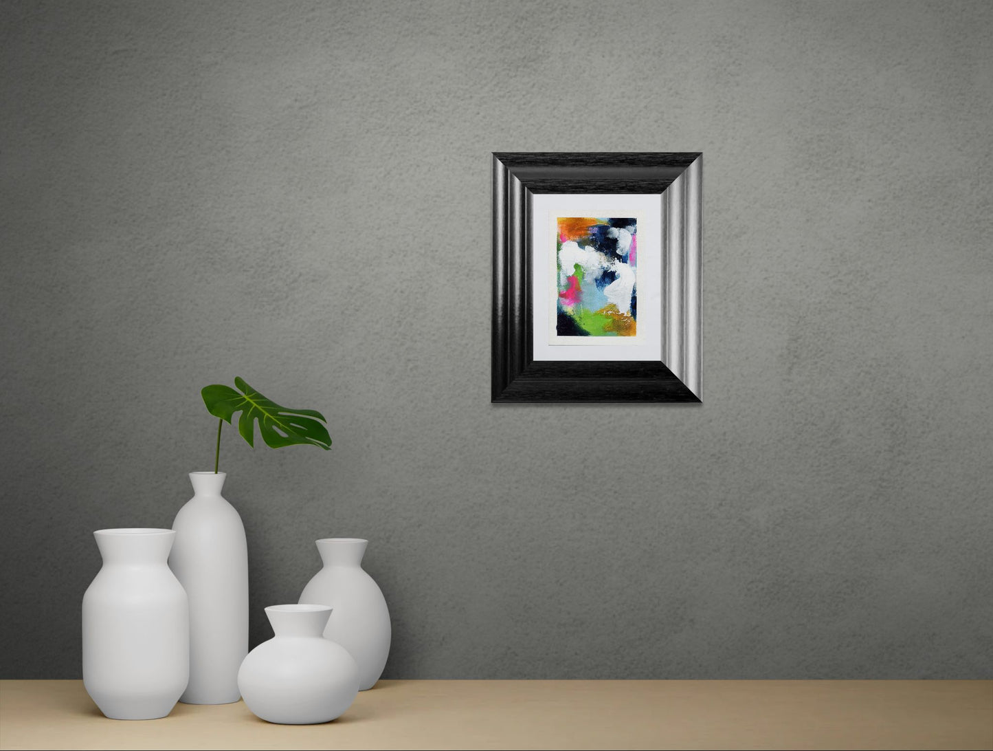 Framed abstract art piece on a gray wall with white vases and a plant below.