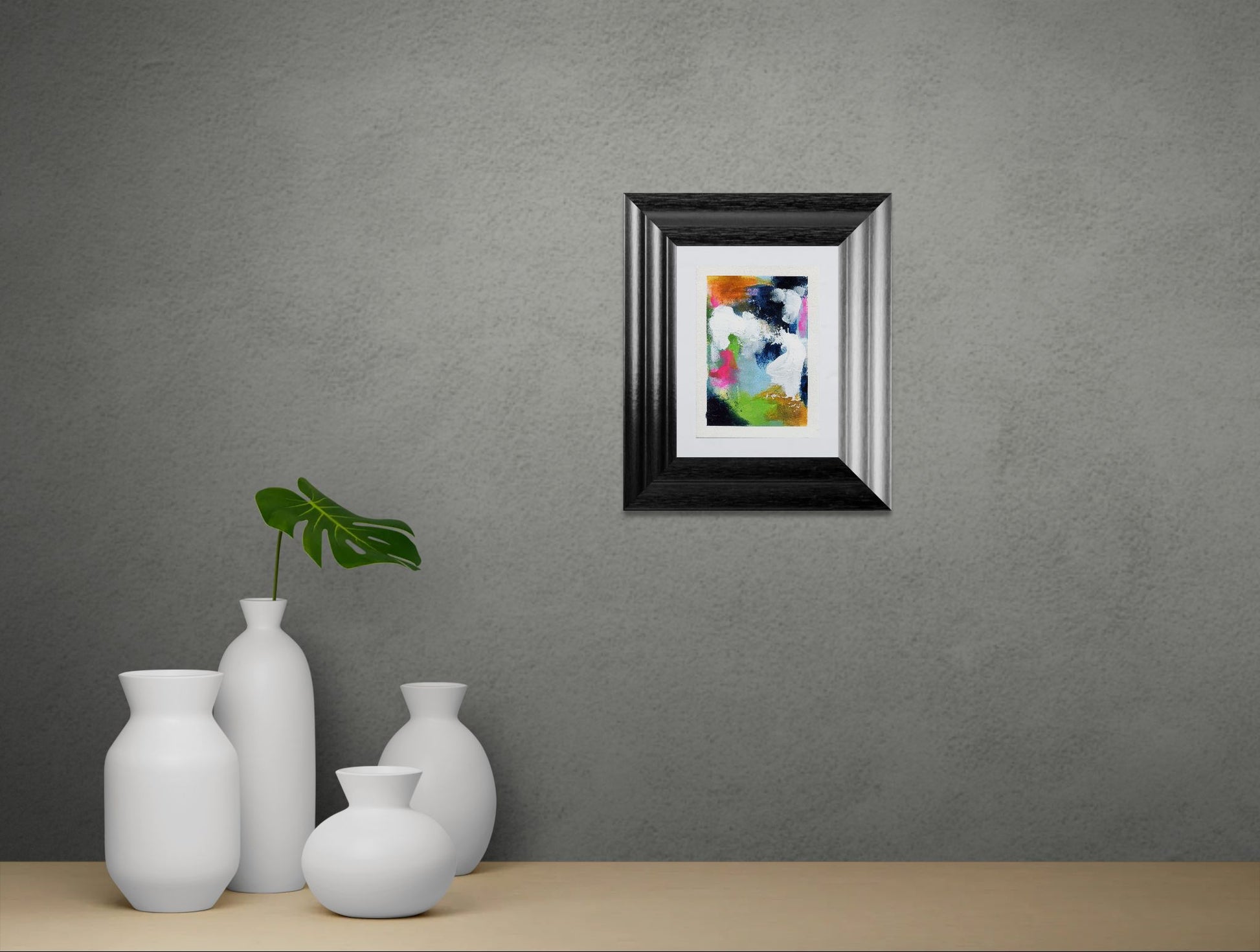 Framed abstract art piece on a gray wall with white vases and a plant below.