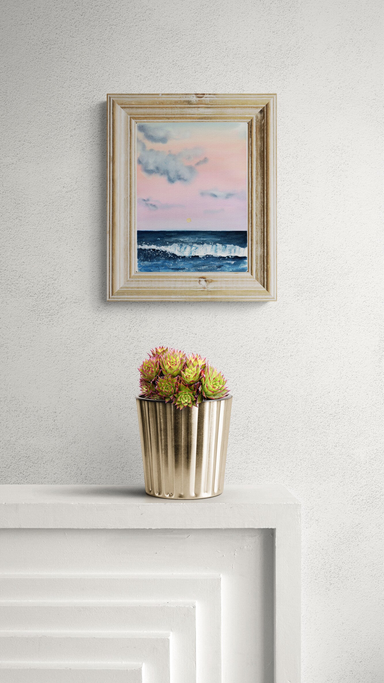 Framed artwork of a seascape with a potted plant below on a white wall.