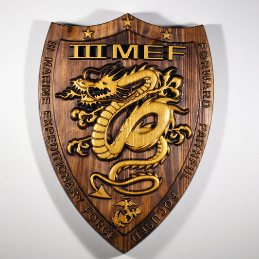 A 3D wood carving plaque featuring the USMC I Marine Expeditionary Force logo with a gold and bronze colored dragon design and the text '1 MEF FORWARD'. The plaque has a brown mahogany finish.