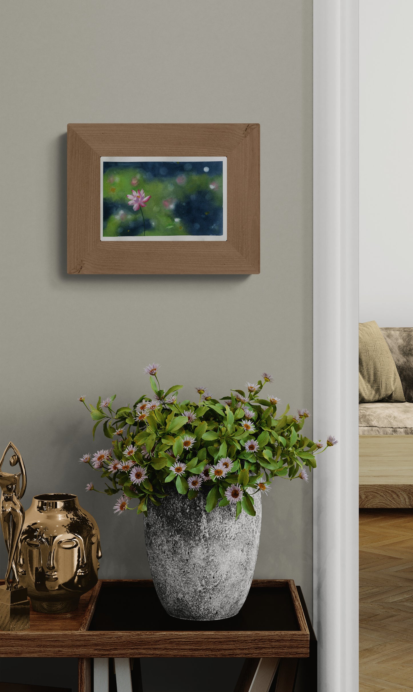 A framed art print depicting a pond scene with green lily pads and pink flowers, styled on a wall above a potted plant on a wooden console table.