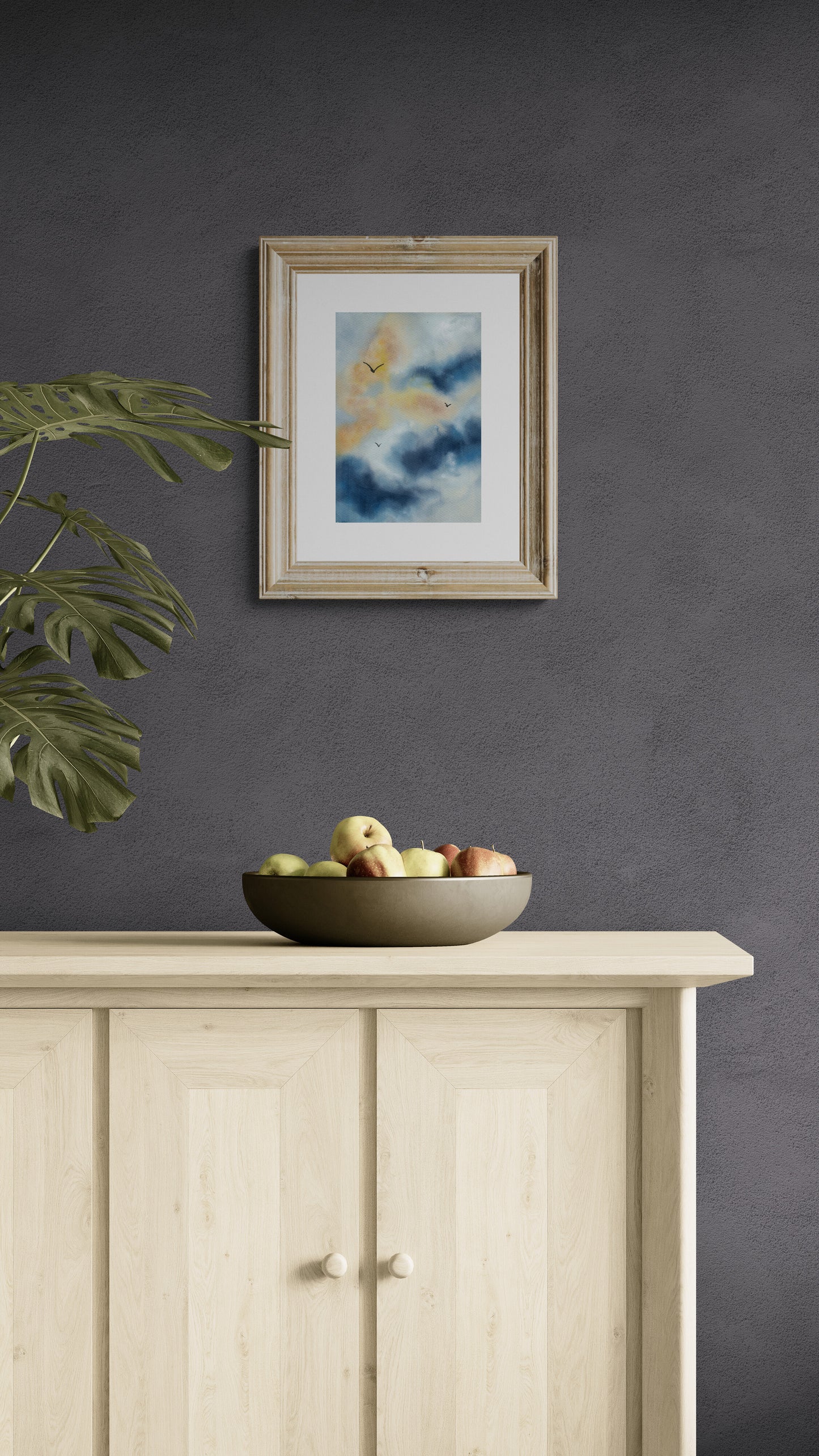 Framed artwork on a dark wall above a wooden cabinet with a bowl of fruit.