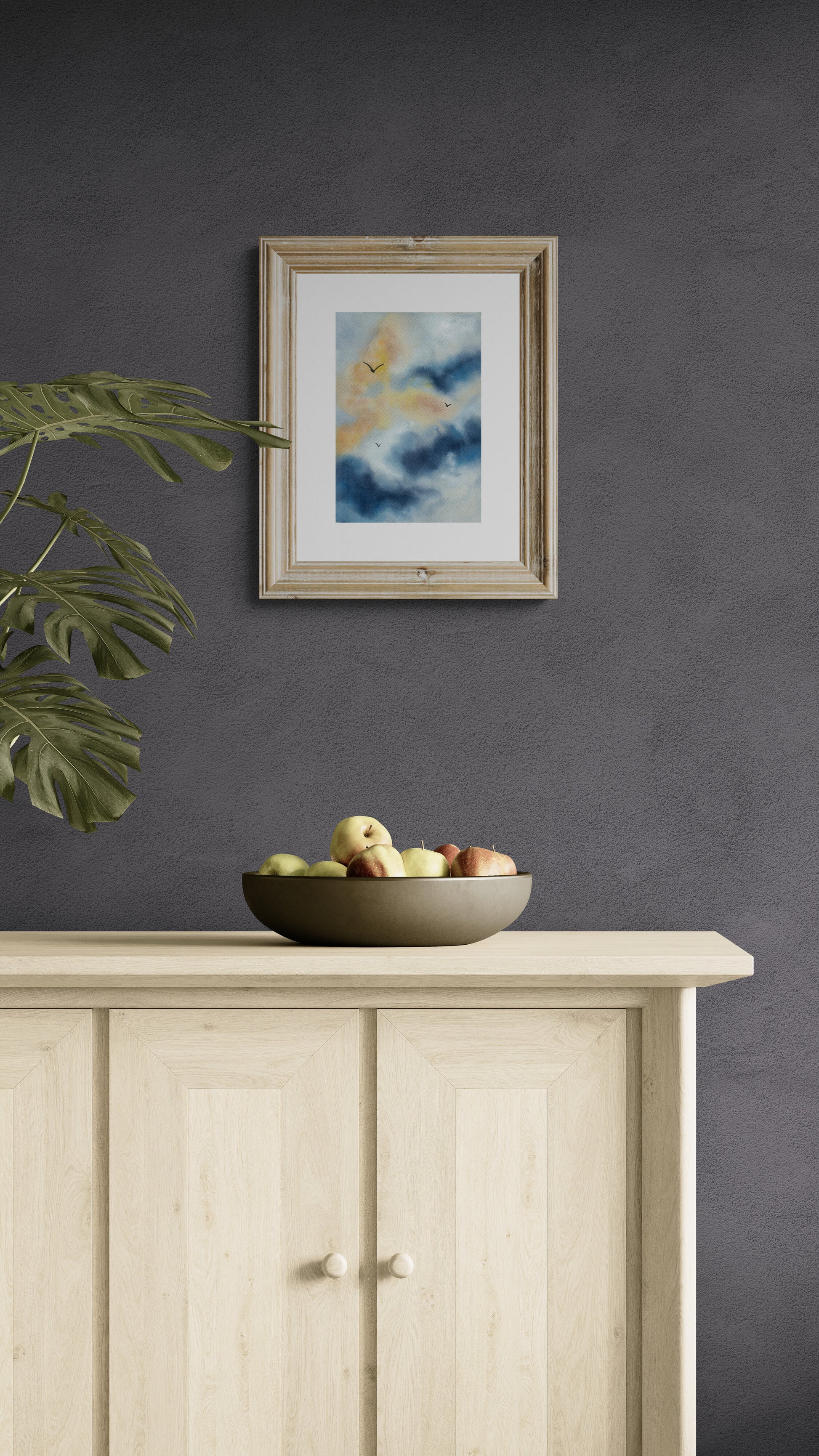 Framed artwork on a dark wall above a wooden cabinet with a bowl of fruit.
