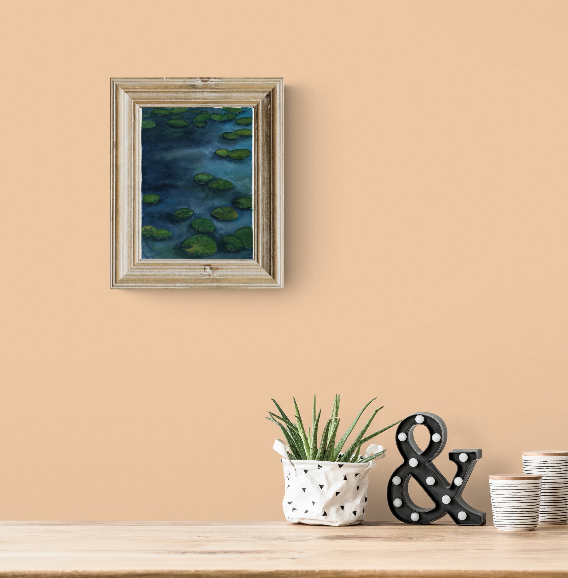 A framed art print depicting green lily pads on a dark blue pond, hung on a warm beige wall above a wooden table styled with a potted succulent, a black ampersand decor piece, and stacked plates.