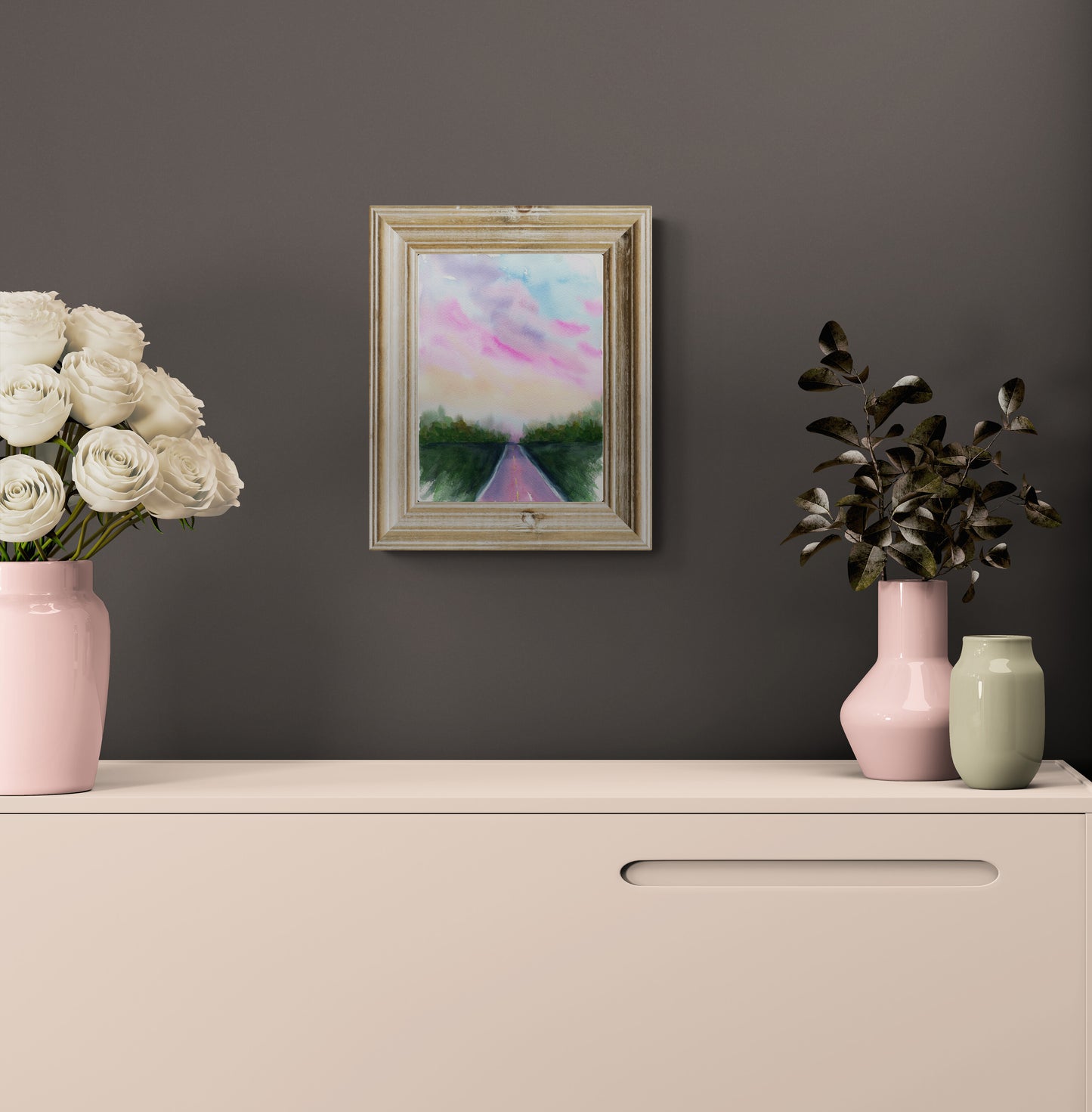 A framed landscape painting hanging on a dark gray wall above a cream-colored cabinet. The painting depicts a pink and blue sky over a green path.