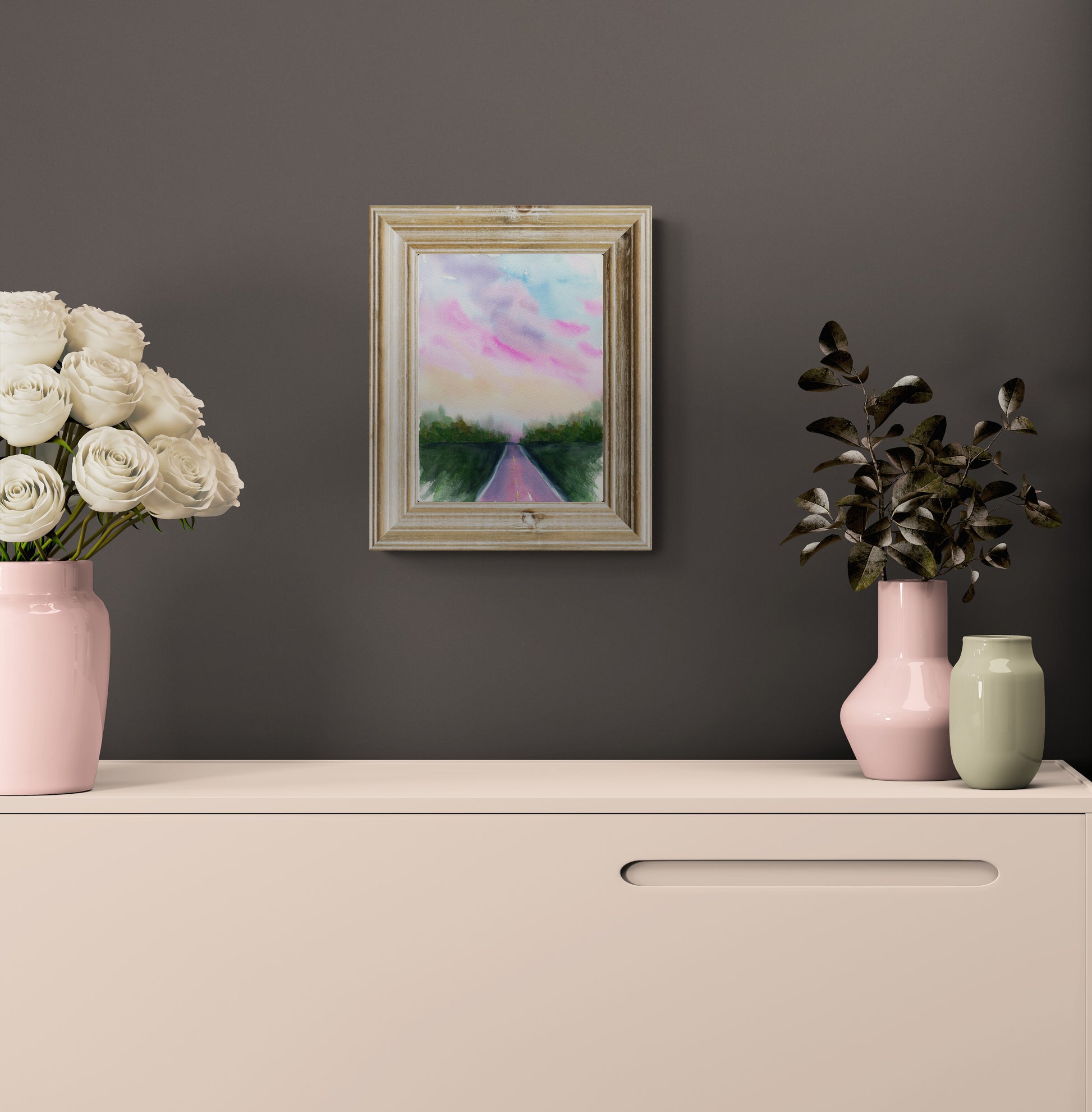 A framed landscape painting hanging on a dark gray wall above a cream-colored cabinet. The painting depicts a pink and blue sky over a green path.