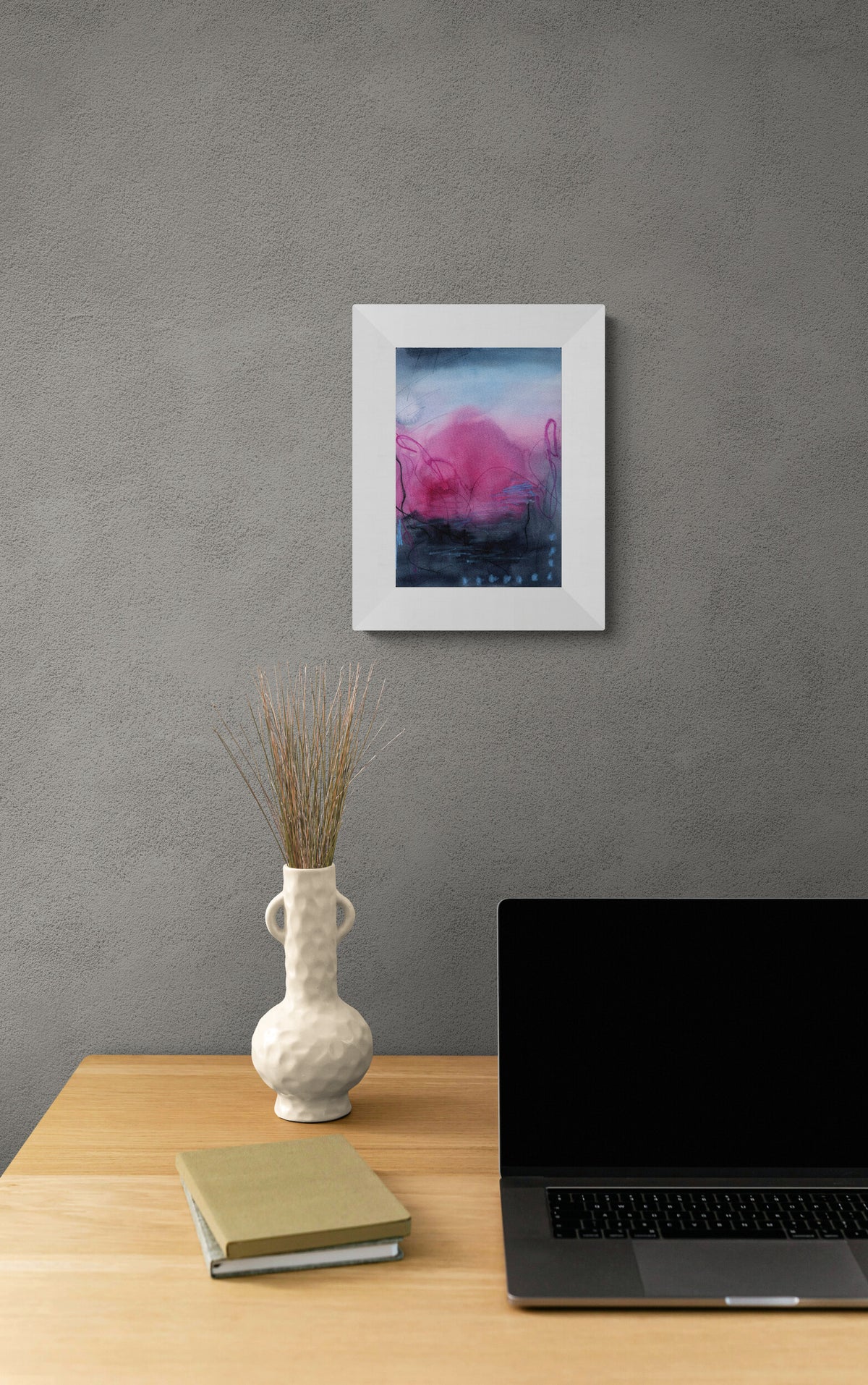 A framed abstract art print featuring pink, purple, and blue tones, displayed on a textured gray wall above a wooden desk with a laptop, vase, and books.