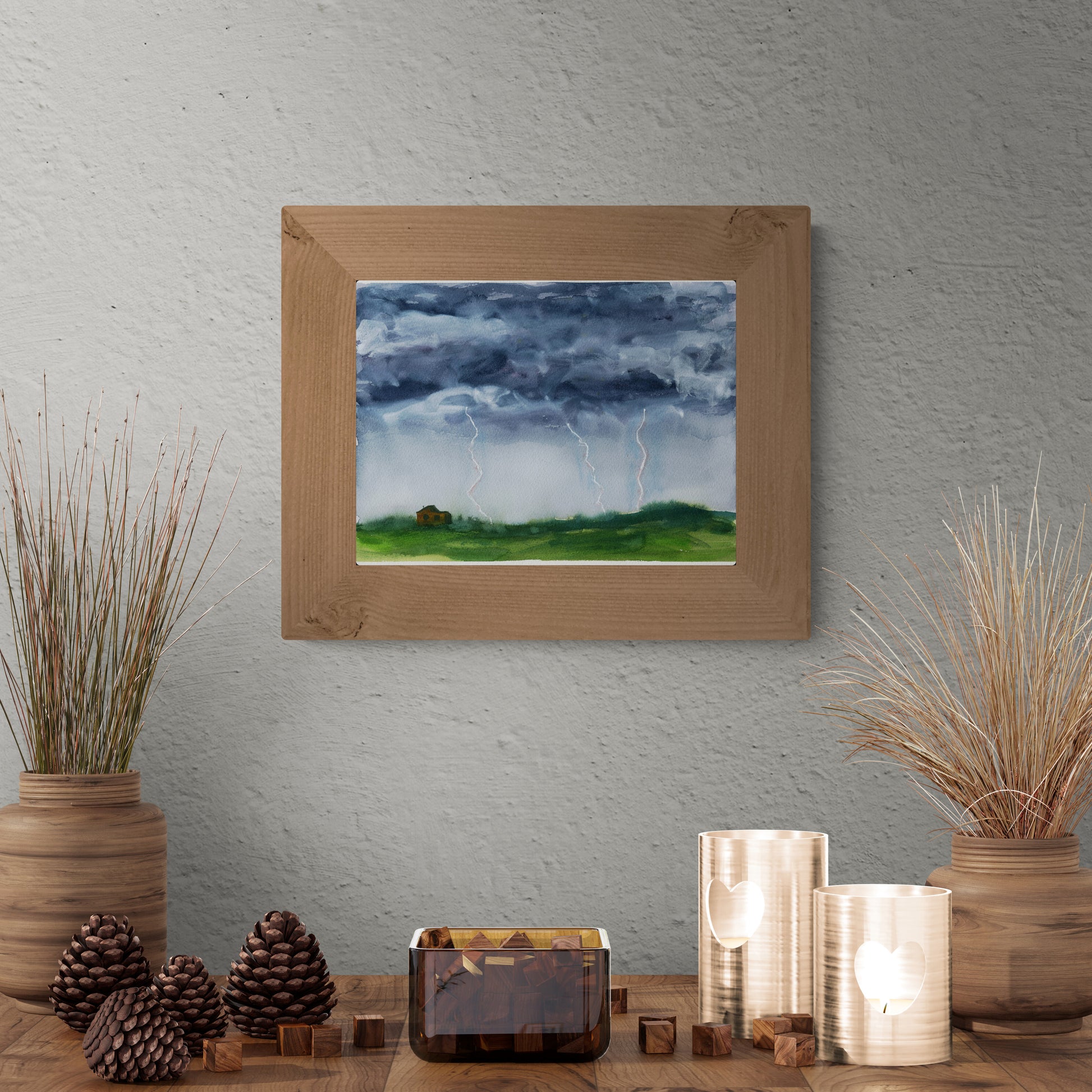 A framed landscape art print depicting a stormy sky with lightning over a green field, displayed on a textured gray wall above a styled wooden table with decorative vases, pinecones, and candles.