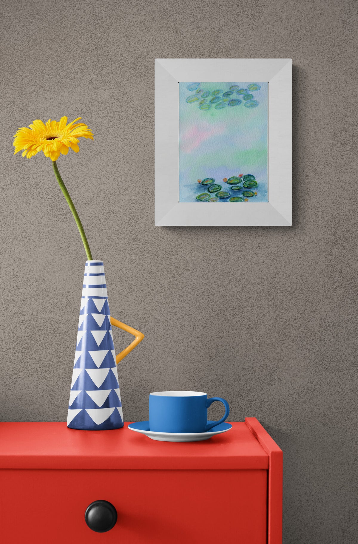 A framed abstract painting of lily pads hangs on a textured gray wall above a bright red side table. The painting features soft pastel shades of green, blue, and pink.