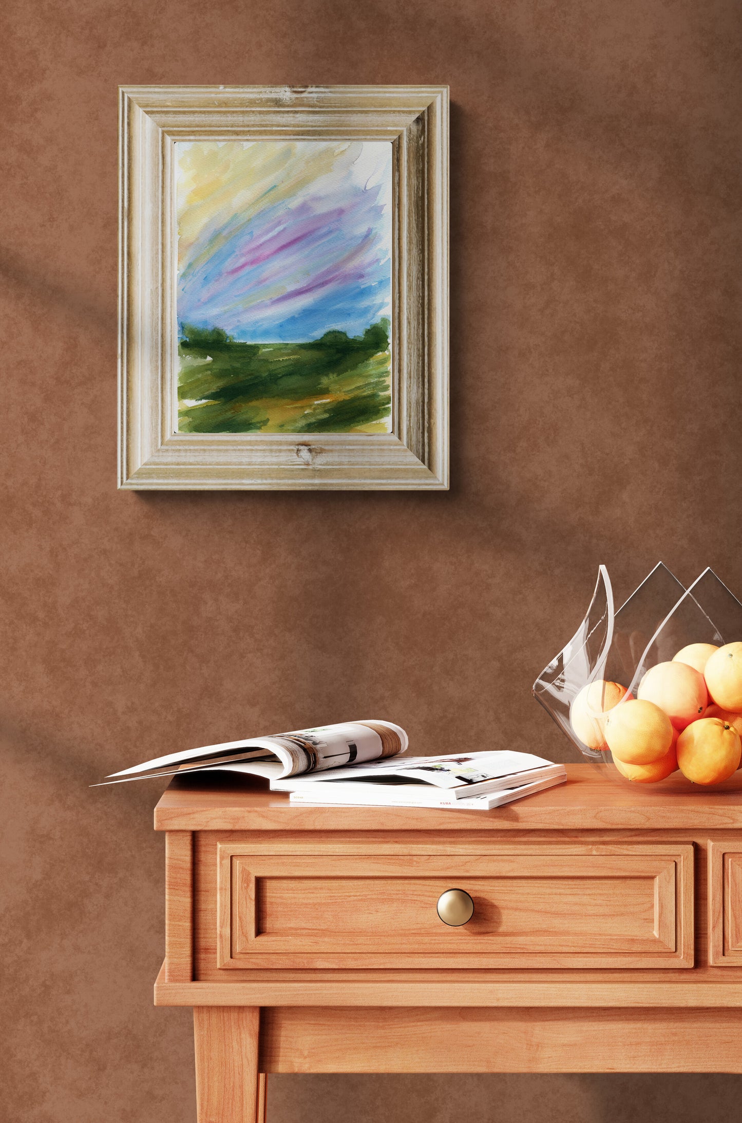 A framed landscape painting with a colorful sky hangs on a textured brown wall above a wooden console table with an open magazine and a glass bowl of oranges.