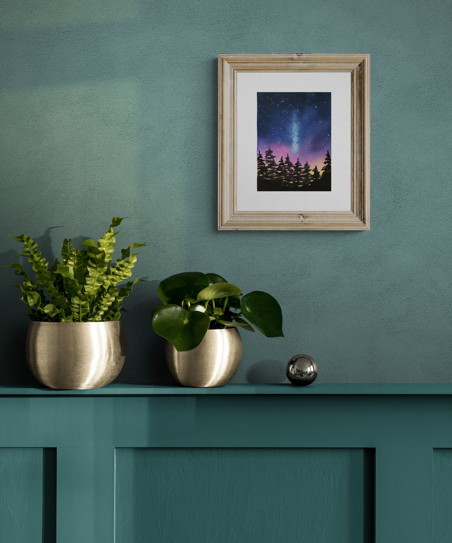 Framed artwork of a starry night sky with trees on a teal wall above a green cabinet with plants.