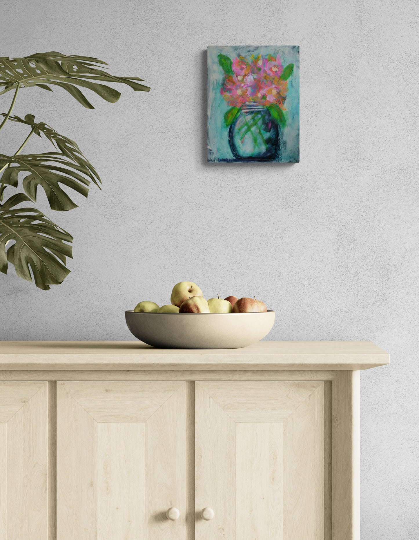A small square canvas painting of pink flowers in a blue vase hangs on a light gray wall above a light wood cabinet, with a bowl of fruit and a large green plant leaf in the foreground.
